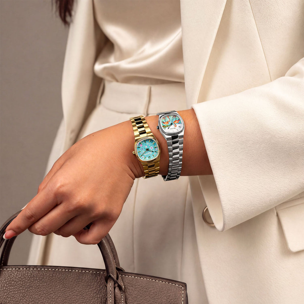 Bulova Super Seville Jardin Bleu — Worn stacked with a silver watch while holding a taupe leather handbag, the custom gold-tone case framing pink and white blossoms on turquoise, customized exclusively by IFL Watches.